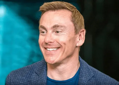 A man with blonde hair and a blue shirt smiles at the camera.