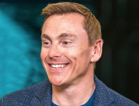 A man with a blue jacket and a blue shirt smiling.